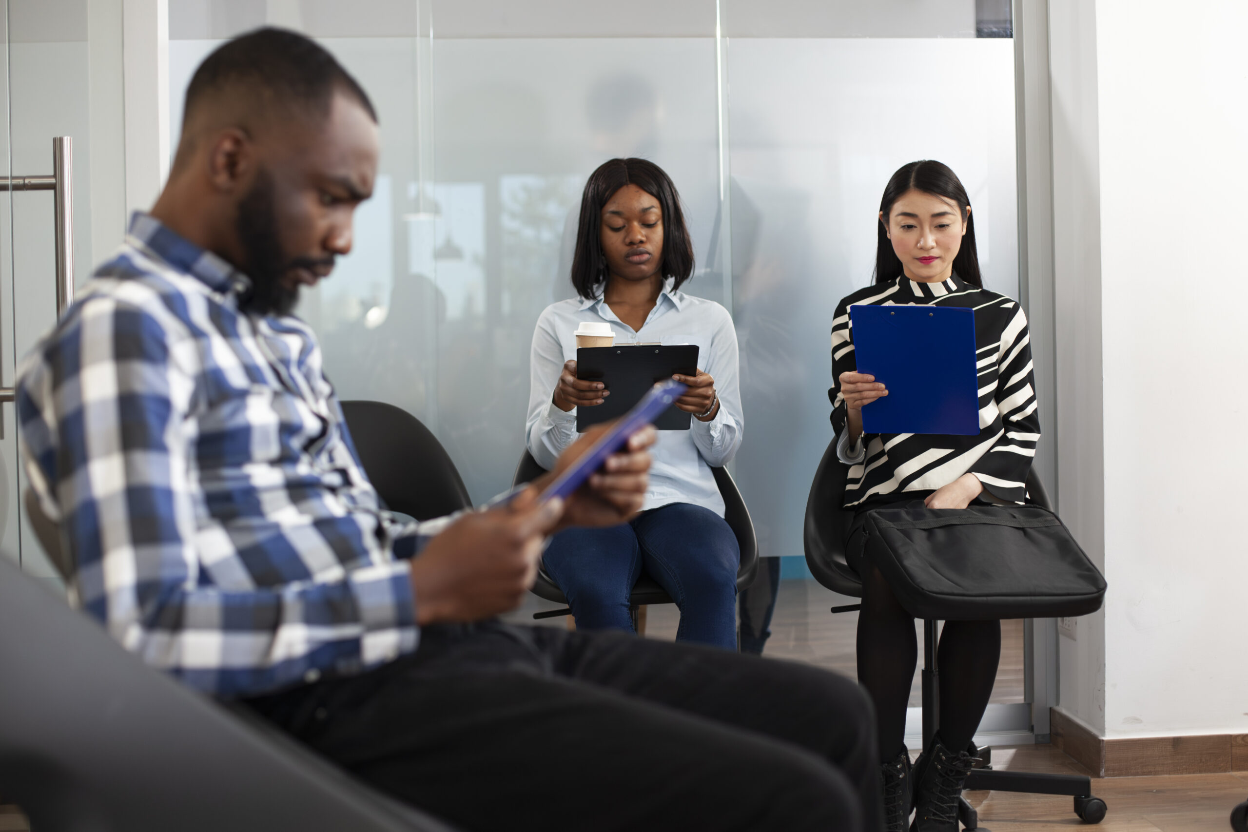 Young applicants waiting in office lobby Young women seated together in office lobby, reading company regulations and waiting for interview. Multiethnic male and female job applicants sitting in queue and preparing for employment meeting.
